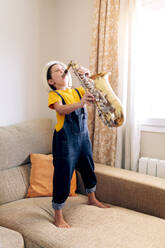 Barfüßiges Kind mit geschlossenen Augen, das Saxophon spielt, während es tagsüber zu Hause auf der Couch steht - ADSF29965