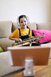 Mindful child in headphones and saxophone on couch recording video on cellphone at home - ADSF29967