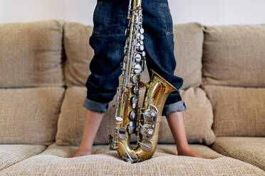 Cropped anonymous thoughtful barefoot child in hat with saxophone standing on sofa against window at home - ADSF29969