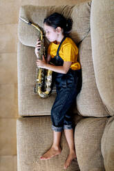 Von oben Seitenansicht des barfüßigen Kindes mit Saxophon, das auf der Couch im Hauszimmer schläft - ADSF29970