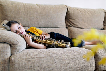 Entzückendes Kind mit Saxophon, das auf der Couch im Haus schläft - ADSF29971
