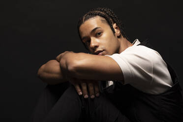 Side view of confident young ethnic hipster male model with braided hair wearing white shirt with black clothes looking at camera while sitting in dark studio - ADSF29980