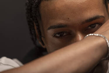 Headshot of serious young African American male with braided hair and bracelet on wrist looking at camera thoughtfully - ADSF29981