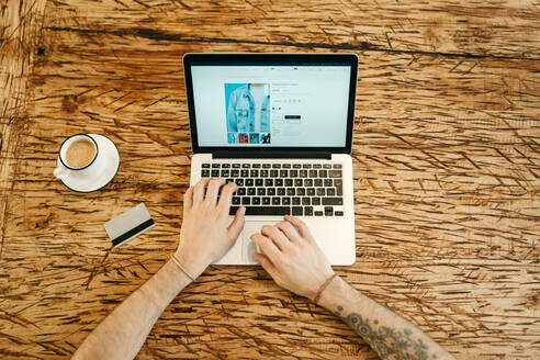 Draufsicht auf einen anonymen Mann, der beim Online-Shopping auf einer Website Kleidung auswählt, während er ein Netbook benutzt und mit einer Plastikkarte in einem Café am Tisch sitzt - ADSF29984