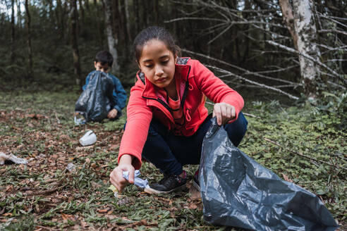 Ethnische Freiwillige mit Plastiksäcken, die im Sommerwald bei Tageslicht Müll vom Gelände gegen Bäume sammeln - ADSF30010