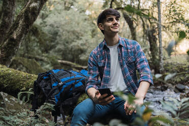 Seitenansicht eines zufriedenen männlichen Wanderers mit Rucksack, der in der Nähe eines Waldsees sitzt und auf seinem Mobiltelefon surft - ADSF30012
