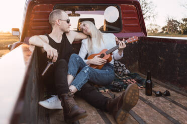 Loving couple sitting in trunk of vintage pickup car and playing ukulele while chilling in countryside in summer evening and looking at each other - ADSF30015