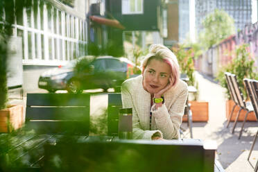 Ungezwungene Frau mit gefärbten Haaren, die an einem Tisch in einem Straßencafé sitzt, sich auf eine Hand stützt und in Träume blickt - ADSF30017