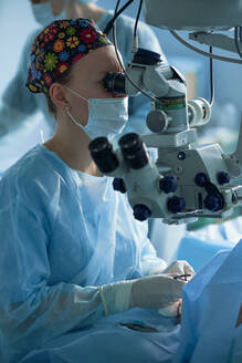 Erwachsener weiblicher Arzt in steriler Maske und ornamentaler medizinischer Kappe, der durch ein Operationsmikroskop gegen einen Mitarbeiter im Krankenhaus blickt - ADSF30024