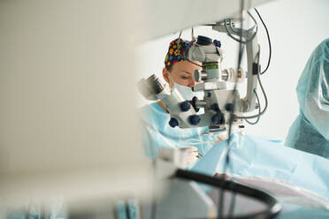 Attentive female doctor in surgical uniform and sterile mask looking through microscope while operating eye of unrecognizable patient in hospital - ADSF30030