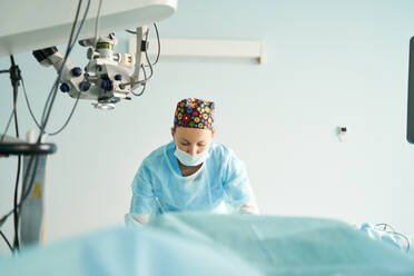 Attentive adult female medic in disposable mask and ornamental cap working in clinic with surgical microscope - ADSF30032