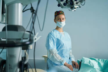 Adult female doctor in surgical uniform and sterile mask looking at camera while sitting in clinic - ADSF30033