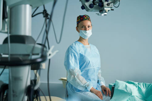 Erwachsene Ärztin in chirurgischer Uniform und steriler Maske schaut in die Kamera, während sie in einer Klinik sitzt - ADSF30033