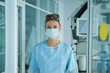 Fröhliche erwachsene Sanitäterin mit steriler Maske und Zierkappe schaut in die Kamera im Krankenhaus - ADSF30034