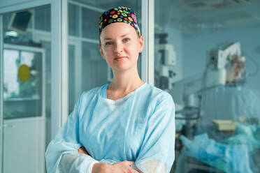 Self assured adult female doctor with folded arms in ornamental medical cap looking at camera against glass wall in hospital - ADSF30035