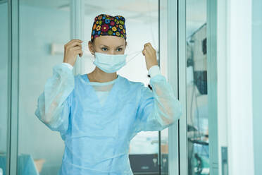 Erwachsene Ärztin in chirurgischer Uniform und ornamentaler medizinischer Kappe, die eine Einwegmaske aufsetzt, während sie im Krankenhaus nach vorne schaut - ADSF30037