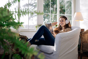 Content female in wireless headset browsing internet on tablet while listening to song in armchair at home - ADSF30045