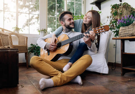 Fröhlicher tätowierter männlicher Musiker spielt Gitarre in der Nähe der zufriedenen weiblichen Geliebten, während sie sich gegenseitig im Sessel im Hauszimmer ansehen - ADSF30052