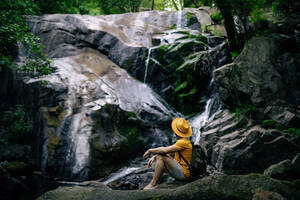Seitenansicht eines nicht erkennbaren männlichen Wanderers, der auf einem Felsen sitzt und einen Wasserfall im Wald bewundert - ADSF30053