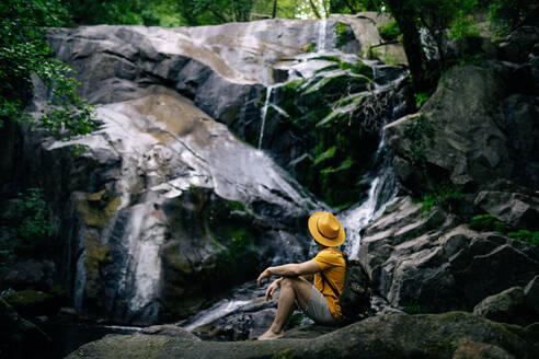 Side view of unrecognizable male hiker sitting on boulder and admiring waterfall in forest - ADSF30053