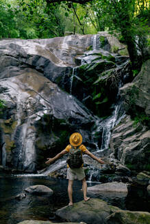 Rückenansicht eines nicht erkennbaren männlichen Wanderers, der auf einem Felsen steht und mit offenen Armen einen Wasserfall im Wald bewundert - ADSF30054