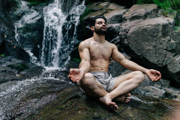 Friedlicher Mann ohne Hemd, der in Padmasana mit Mudra-Händen und geschlossenen Augen sitzt, während er Yoga macht und auf einem nassen Felsen in der Nähe eines Wasserfalls meditiert - ADSF30057