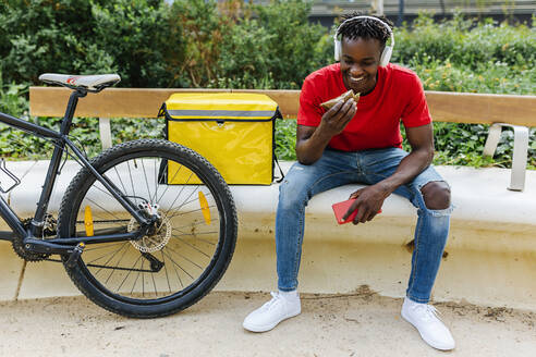 Lächelnder junger Zusteller, der Essen isst, während er mit dem Fahrrad auf einer Bank sitzt - XLGF02290
