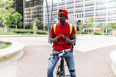 Smiling delivery man holding mobile phone while sitting on bicycle in city - XLGF02308