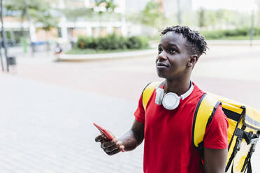 Confused delivery man wearing wireless headphones holding mobile phone - XLGF02311