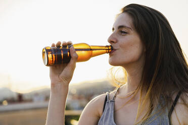 Frau trinkt Bier auf einer Dachterrasse bei Sonnenuntergang - AFVF09179