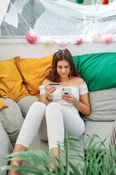 Junge zufriedene Frau, die auf der Couch sitzt und mit einer Plastikkarte für eine Bestellung beim Online-Shopping mit dem Handy bezahlt - ADSF30062