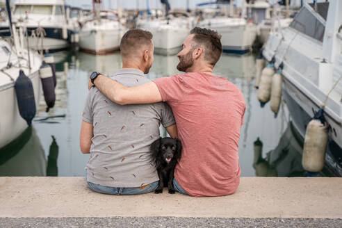 Rückenansicht eines Hundes zwischen einem fröhlichen bärtigen Mann, der einen anonymen homosexuellen Partner umarmt, während er sich unterhält und auf einem Pier im Hafen sitzt - ADSF30133