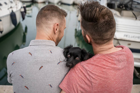 Rückenansicht eines Hundes zwischen einem fröhlichen bärtigen Mann, der einen anonymen homosexuellen Partner umarmt, während er sich unterhält und auf einem Pier im Hafen sitzt - ADSF30134