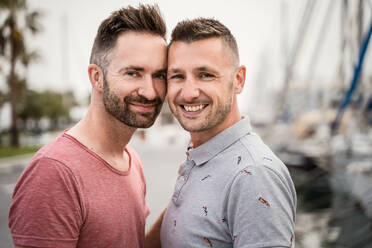 Zufriedenes Paar homosexueller Männer in T-Shirts, die sich umarmen, während sie in der Stadt in die Kamera schauen - ADSF30135
