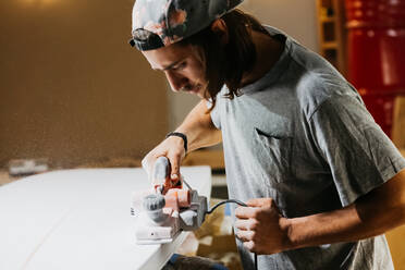 Männlicher Shaper bei der Arbeit mit der elektrischen Hobelmaschine und beim Polieren der Oberfläche eines Surfbretts in der Werkstatt - ADSF30143