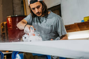 Male shaper using electric planer and polishing surface of surfboard in workshop - ADSF30144