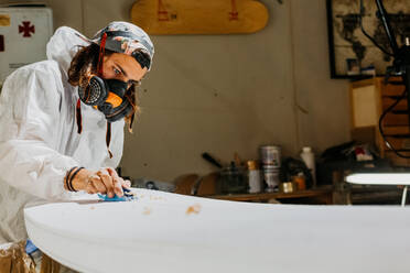 Seitenansicht eines männlichen Shapers in Schutzanzug und Atemschutzmaske, der die Oberfläche eines Surfbretts mit einem Handhobel in einer Werkstatt poliert - ADSF30145