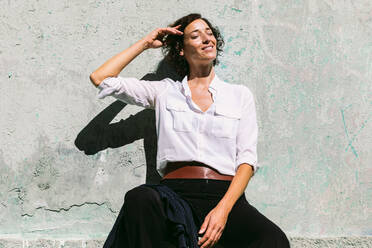 Positive female worker wearing formal clothes sitting near concrete wall and enjoying sunny day while gently touching hair - ADSF30167