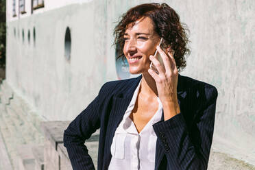 Positiver erwachsener weiblicher leitender Angestellter in stilvollem Outfit, der mit der Hand auf der Taille steht und mit dem Handy in einem sonnigen Tag spricht - ADSF30171