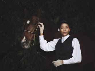 Selbstbewusste afroamerikanische erwachsene Dame in eleganter Kleidung und Hut, die mit einem braunen Pferd steht und in die Kamera blickt, in der Nähe von Bäumen bei Tageslicht - ADSF30208