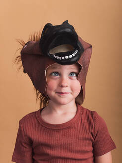 Child in t shirt and horse mask on head looking at camera on beige background - ADSF30216