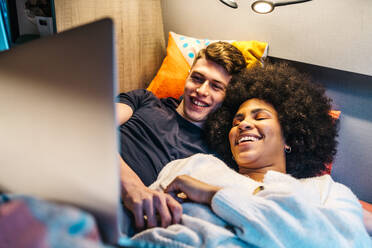 Glückliche romantische multirassische Paar kuscheln und genießen Zeit zusammen, während auf dem Bett liegen und beobachten Film auf dem Laptop während der Reise im Wohnmobil van - ADSF30223