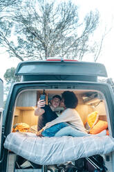 Young multiracial couple in love taking selfie on smartphone while hugging and kissing happily inside camper van during romantic journey together - ADSF30224