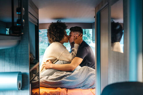 Side view of young multiracial couple in love kissing each other while sitting on bed inside camper van parked in nature during romantic holidays together in summertime - ADSF30226
