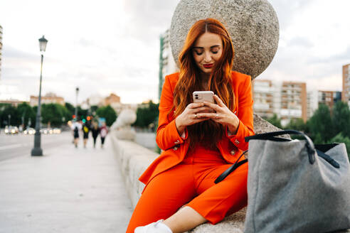 Glücklich stilvolle Frau mit roten Haaren und in lebendigen orange Anzug sitzt auf Stein Grenze in der Stadt und Messaging auf Handy - ADSF30229