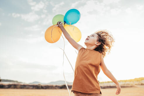 Lächelndes ethnisches Kind mit lockigem Haar, das mit bunten Luftballons im Sommerfeld spielt und nach oben schaut - ADSF30242