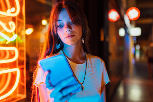 Interessierte junge Frau mit Ohrringen surft mit ihrem Handy im Internet vor leuchtenden Neonröhren in der Stadt - ADSF30252