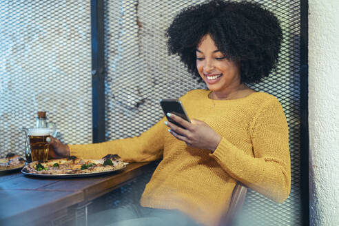 Lächelnde Afro-Frau, die ein Mobiltelefon benutzt, während sie am Tisch etwas trinkt - JSRF01585