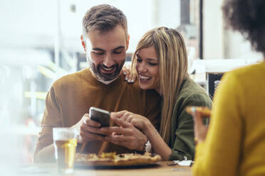 Happy male and female friends sharing smart phone in restaurant - JSRF01604
