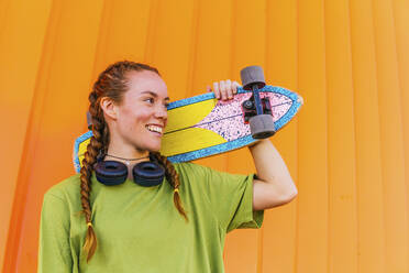 Glückliche junge Frau mit Skateboard vor einer Wand - MGRF00426